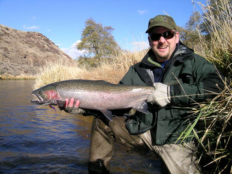 Jim kneeling at the side of a river, holding a large trout and smiling at the camera